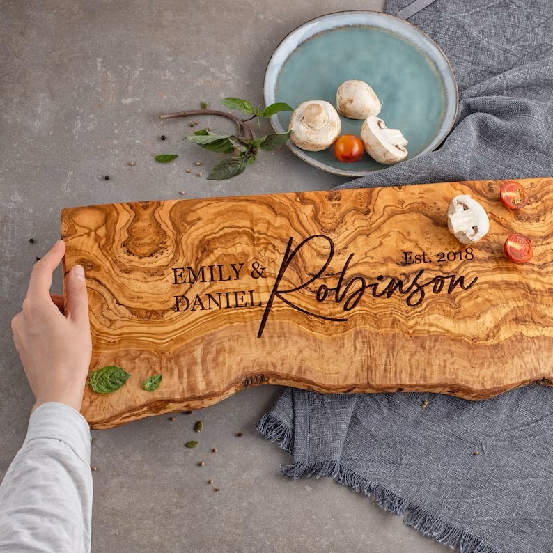 A person holding a personalized Olive Wood Live Edge Board with the couple's first names, last name and date engraved across the board.