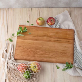 A wooden cutting board on a wooden table with apples and a bag.