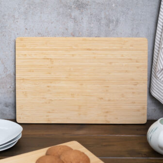A bamboo cutting board on a kitchen counter with dishes and a cloth nearby.