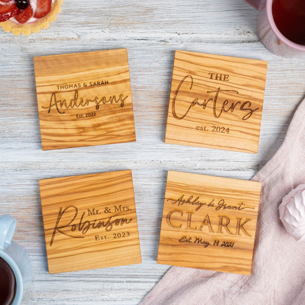 Four personalized wooden coasters with engraved names and establishment dates, displayed on a table with a cup of tea and a pastry in the background.