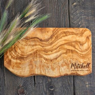 A live edge cutting board made out of olive wood with an engraving in the lower corner laying on top of a wooden table.