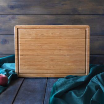A wooden cutting board on a table with a green cloth and red apples on the side.