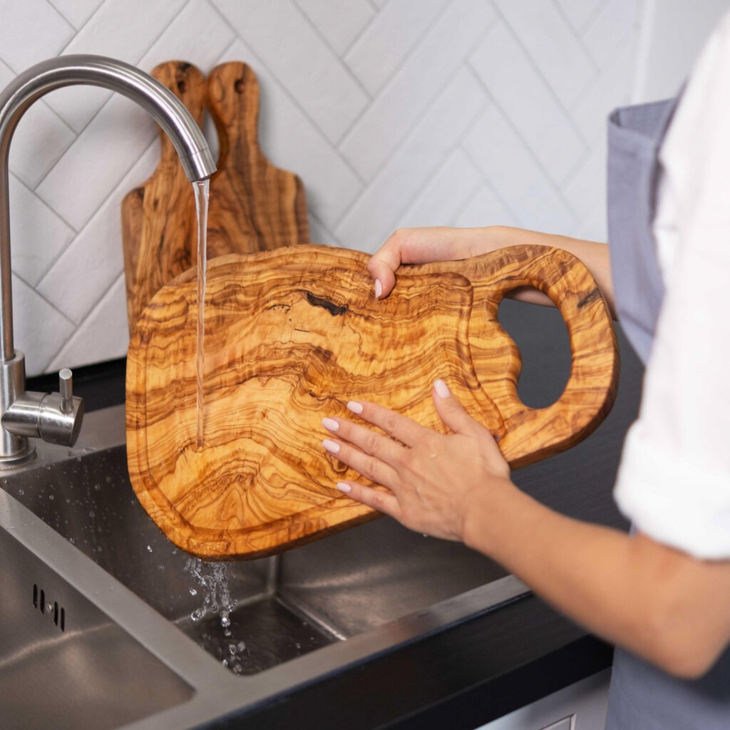 A woman is washing a wooden cutting board in a kitchen sink.