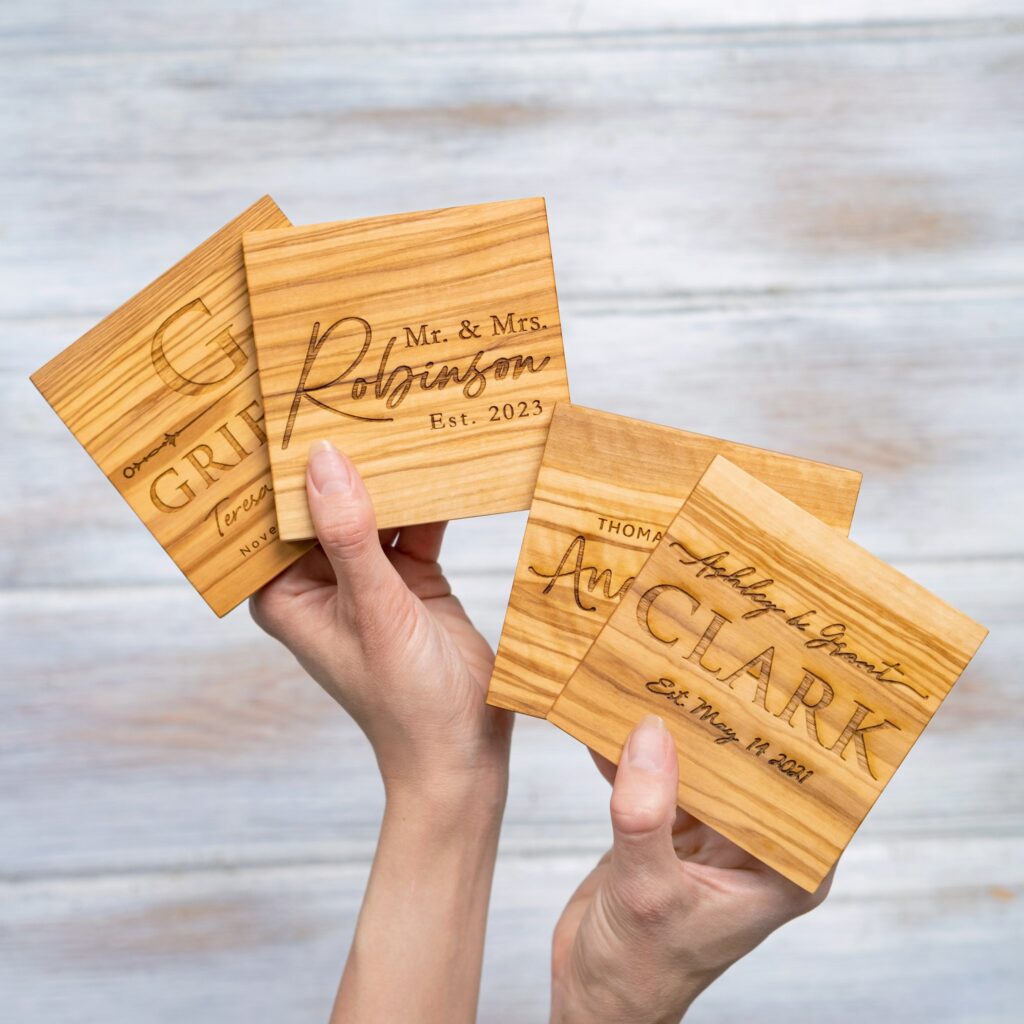 Hands holding personalized wooden coasters with engraved names and wedding dates, set against a wooden background.