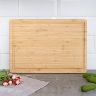Bamboo cutting board on a kitchen countertop with radishes and cucumbers nearby.