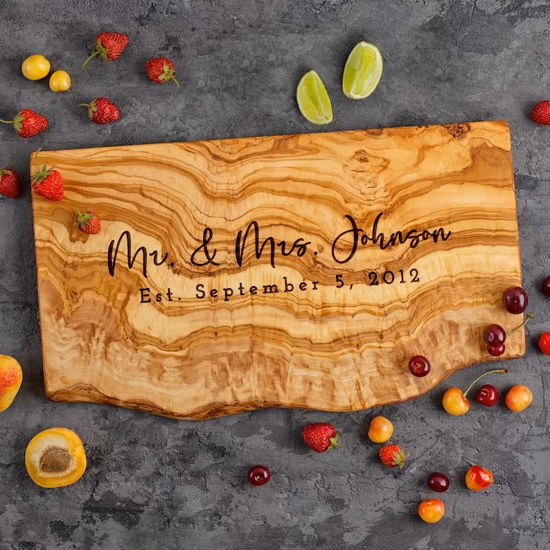 Personalized olive wood live edge cheese board laying on top of a kitchen counter surrounded by pieces of fruit.