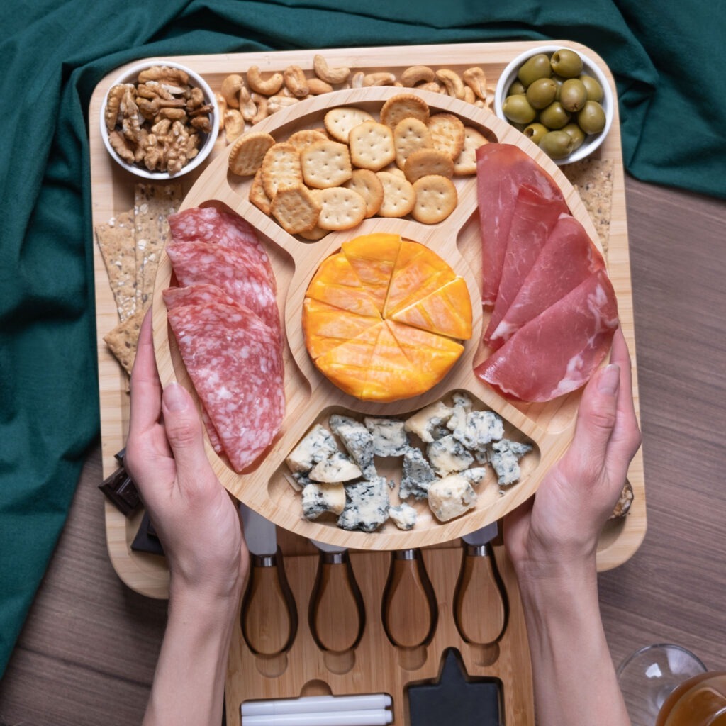 A person holding a wooden cheese board with a selection of cheeses, cured meats, crackers, and nuts.