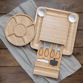 A wooden cheese board set with utensils on a wooden table beside a round wooden serving platter.