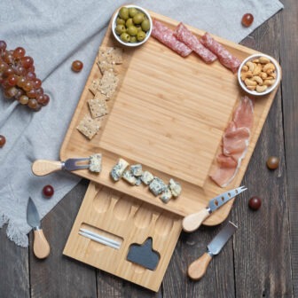 A wooden cheese board with assorted cheeses, cold cuts, olives, grapes, crackers, and nuts, along with cheese knives.