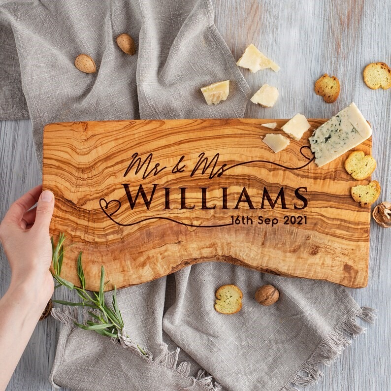 A person holding a live edge olive wood cutting board with the engraved Mr. & Mrs., last name and date across the board.
