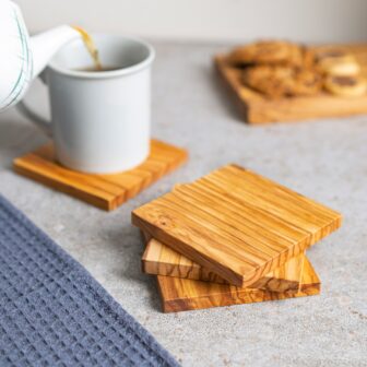 A stack of wooden coasters on a kitchen counter with a mug of tea being poured and cookies in the background.