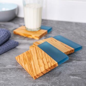 Three wooden coasters with blue silicone accents on a gray countertop, next to a glass of milk and a blue bowl.