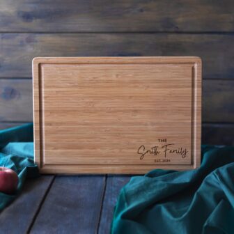 Wooden cutting board with personalized engraving on a table with a teal cloth and apples in the background.
