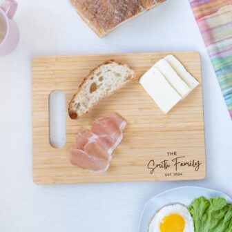 A wooden cutting board with bread, cheese, and prosciutto, personalized with "the smith family est. 2024" engraving.
