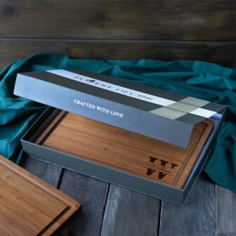 A wooden serving tray with personalization, presented in an open gift box that reads "crafted with love" on a wood surface with a teal cloth.