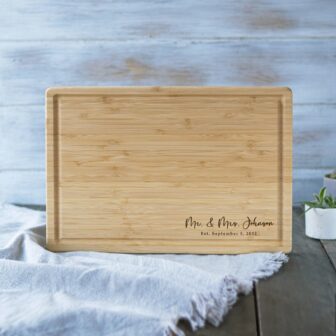 A personalized bamboo cutting board with "mr. & mrs. johnson est. september 5, 2012" engraved on it, placed on a table with a white cloth.