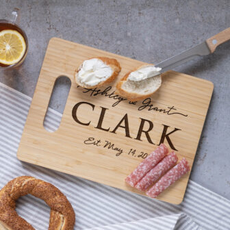 Personalized cutting board with a couple's name, accompanied by a spread of bread, cream cheese, salami, and pretzel with a cup of tea on the side.