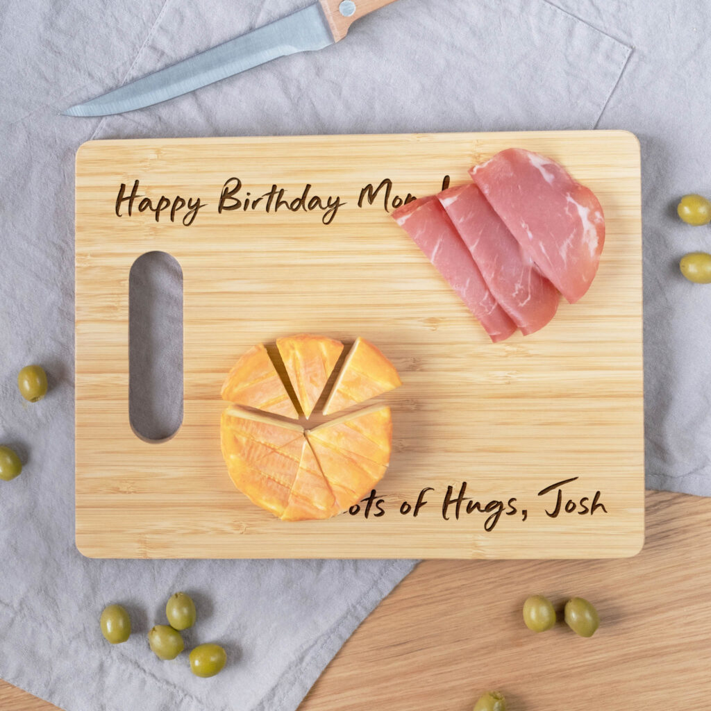 A wooden cutting board with a birthday message, surrounded by slices of cheese and ham, and green olives.