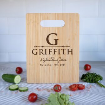 Personalized bamboo cutting board with engraved names and date, surrounded by fresh vegetables on a kitchen countertop.