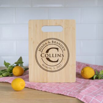 Personalized wooden cutting board with names "joanna & branden collins" and the date "march 3, 2017" engraved, placed on a kitchen counter with lemons and a pink cloth.