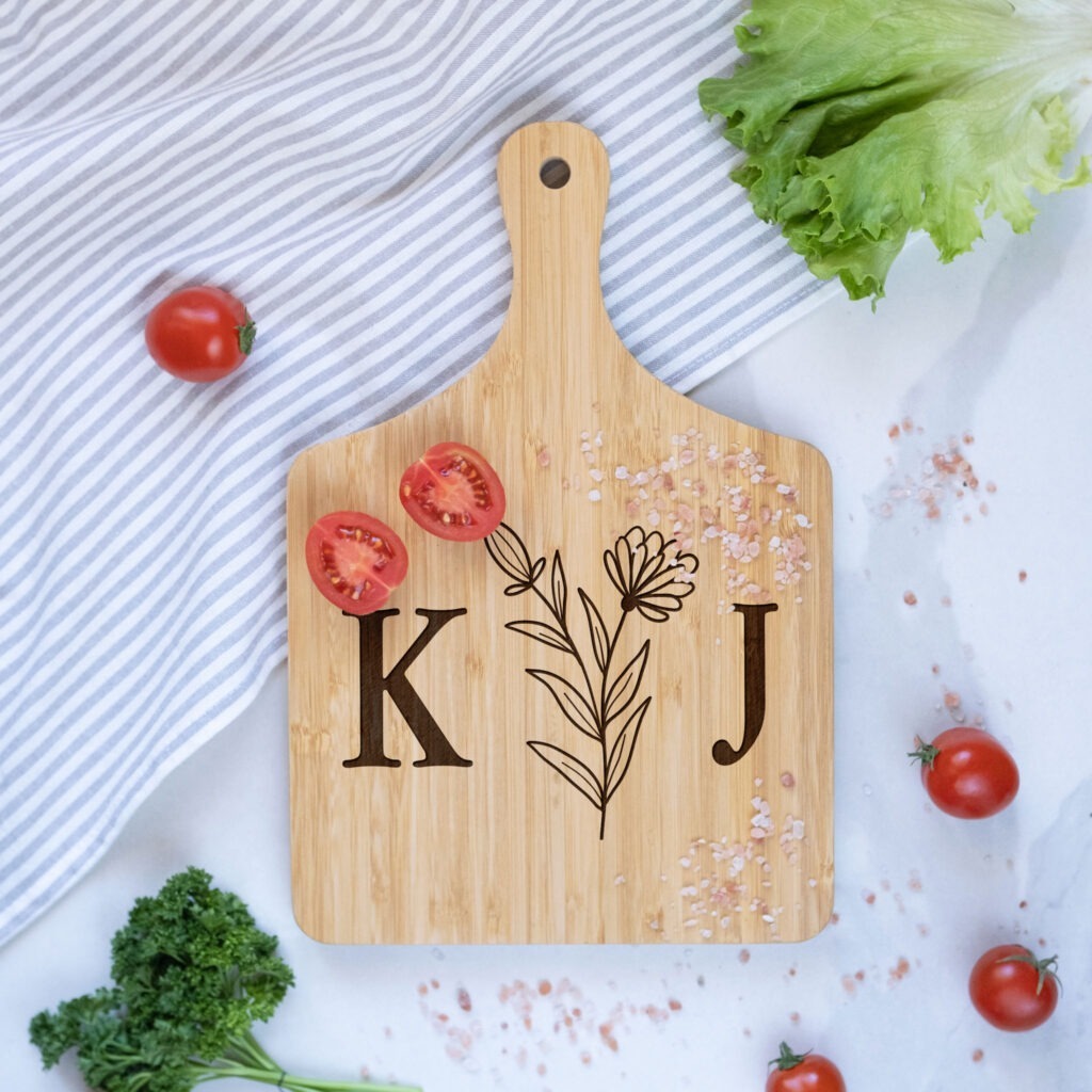 A personalized cutting board with initials "kj" surrounded by fresh vegetables on a kitchen countertop.