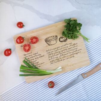 A wooden cutting board with an etched recipe, surrounded by fresh vegetables and a knife on a marble surface.
