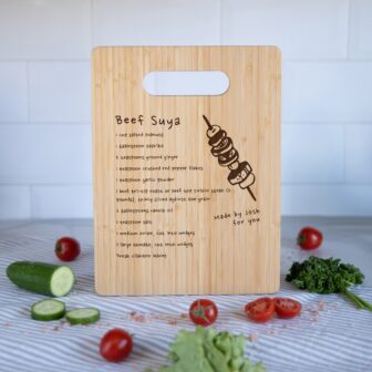 Wooden chopping board with a recipe for beef suya written on it, surrounded by ingredients.