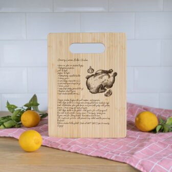 A wooden cutting board with a roasted chicken recipe and illustration, displayed on a countertop with lemons and a pink cloth.