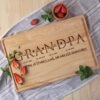 A personalized wooden cutting board with the word "grandpa" engraved, alongside dates and a message about stories, love, and adventures, accompanied by strawberries and mint leaves on a kitchen counter.