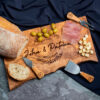 A personalized wooden cheese board with utensils, served with bread, olives, prosciutto, and cheese cubes.