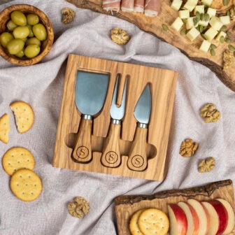 A cheese board set with utensils, surrounded by assorted snacks including crackers, olives, and slices of apple.