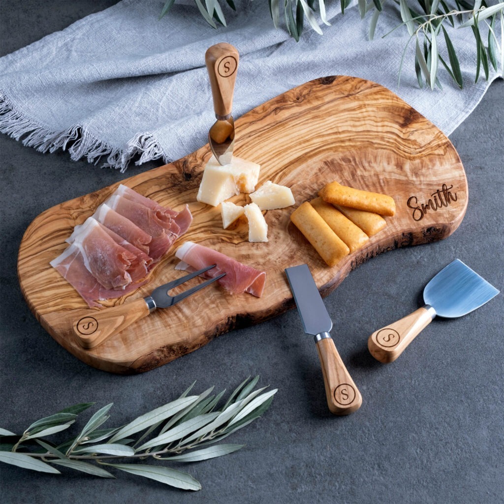 A charcuterie board with cheese, prosciutto, and breadsticks accompanied by serving utensils, garnished with olive branches on a dark surface.