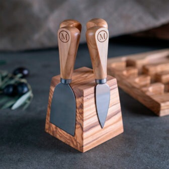 Two cheese knives on a wooden block next to olives.