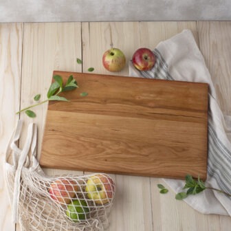 A Wood Cutting Board (Cherry) on a wooden table with apples and a bag.