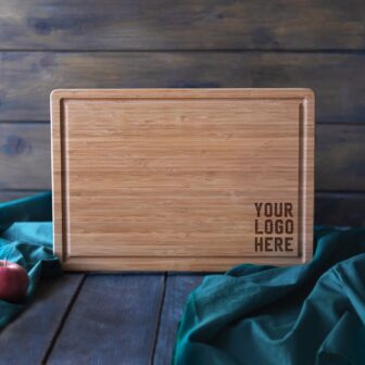 A wooden cutting board with a placeholder text "your logo here" displayed against a blue wooden backdrop with a green cloth and apple in the foreground.