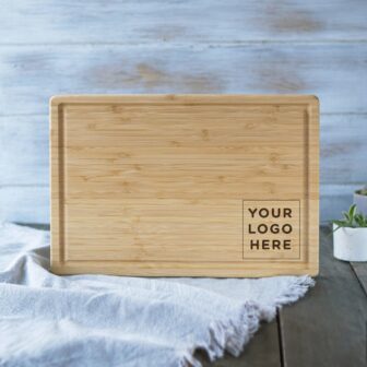 A blank bamboo cutting board with a space for a custom logo displayed on a wooden table with a white cloth.
