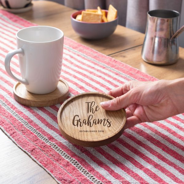 A hand places a round wooden coaster engraved with The Grahams ESTABLISHED 2018 onto a red and white striped table runner next to a white mug.