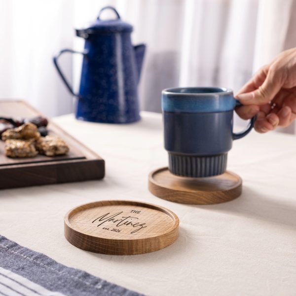 A hand lifts a blue mug from a wooden coaster while an identical coaster engraved with The Martinez est. 2026 sits on the table.