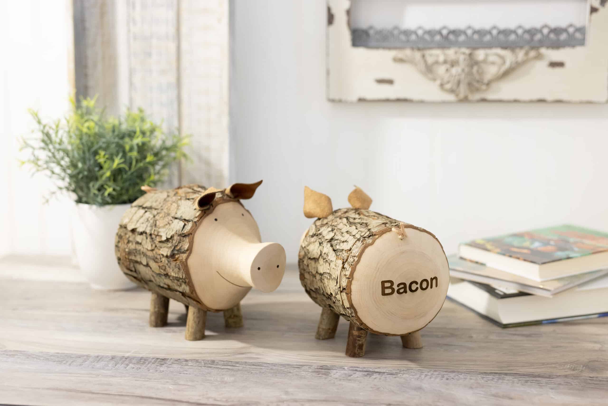 Two decorative wooden pig figurines on a table, labeled "bacon," with a plant and books in the background.