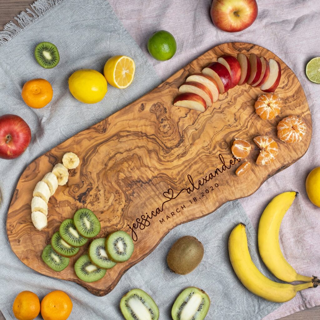 A wooden serving board with an assortment of sliced fruits including kiwis, bananas, apples, and citrus, personalized with engraving.