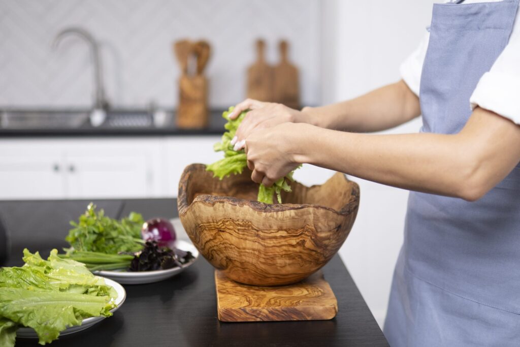 A person in an apron preparing a salad in a wooden bowl on a kitchen counter, surrounded by fresh vegetables.