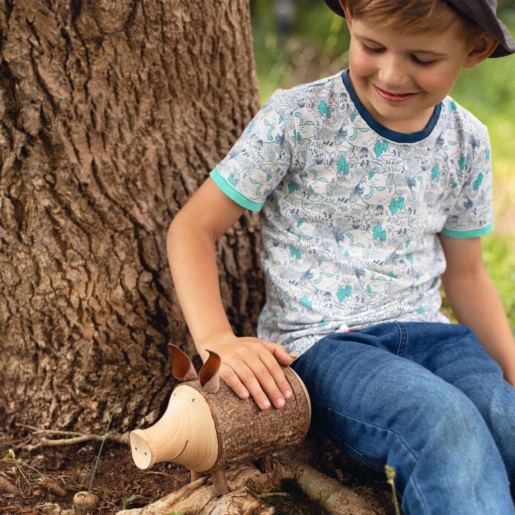 A young boy in a printed shirt and hat smiles while playing with a wooden toy pig next to a large tree.