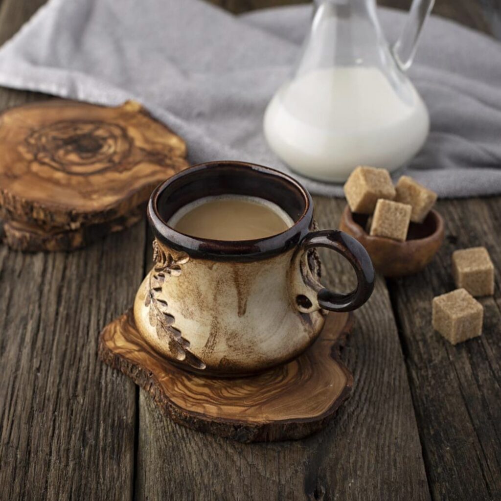 A rustic ceramic mug filled with coffee on a wooden coaster, accompanied by brown sugar cubes and a milk jug on a wooden table.