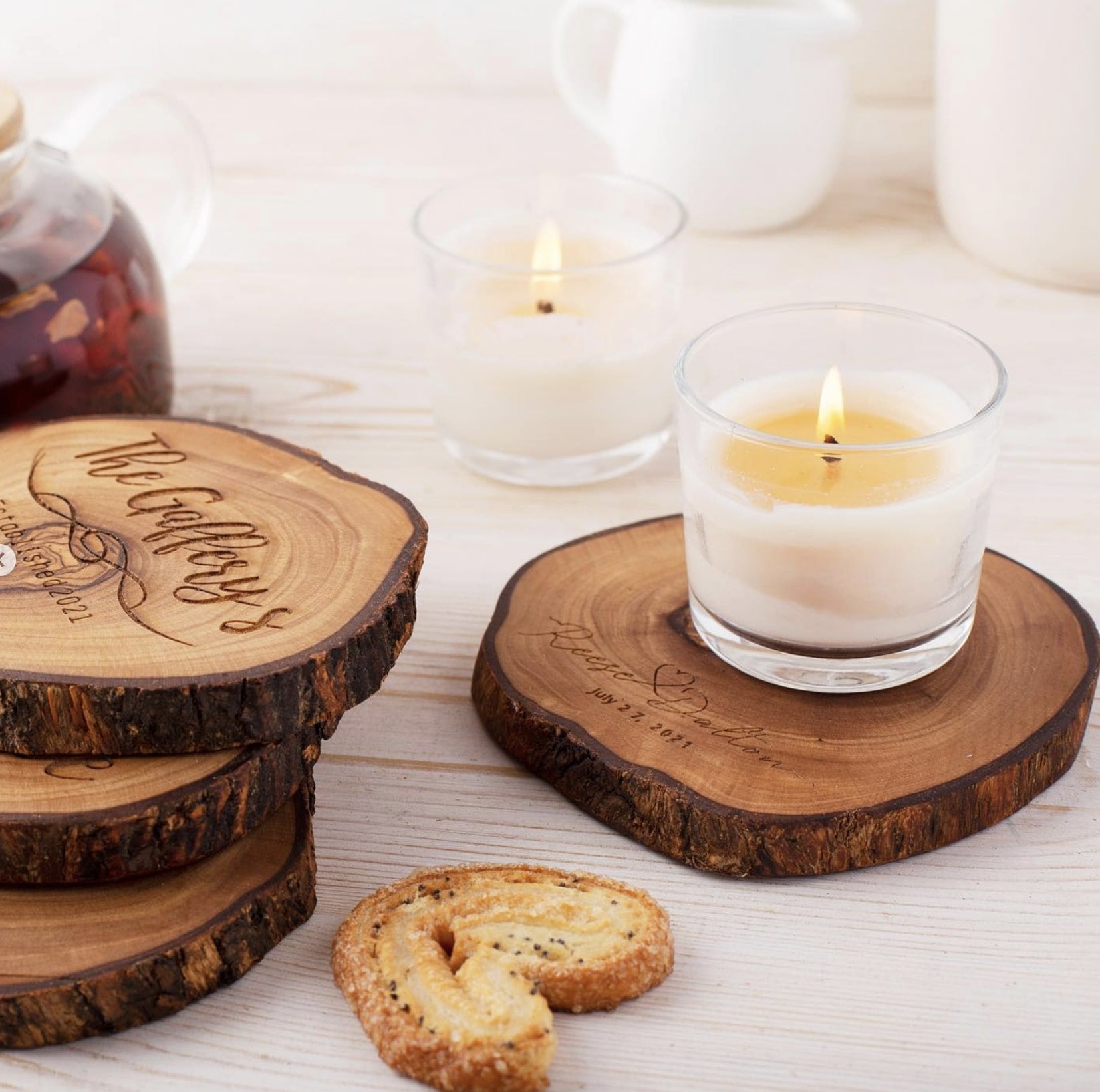 Two candles lit on wooden coasters with engraved messages, next to a teapot, on a white wooden table. a biscuit is also visible near the candles.