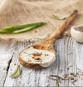 A wooden spoon holds a dollop of creamy dip sprinkled with herbs, set on a rustic table with scattered spices and a burlap cloth in the background.