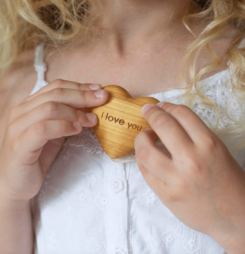 A close-up of a child holding a wooden heart with "i love you" engraved on it, set against a white lacy dress.