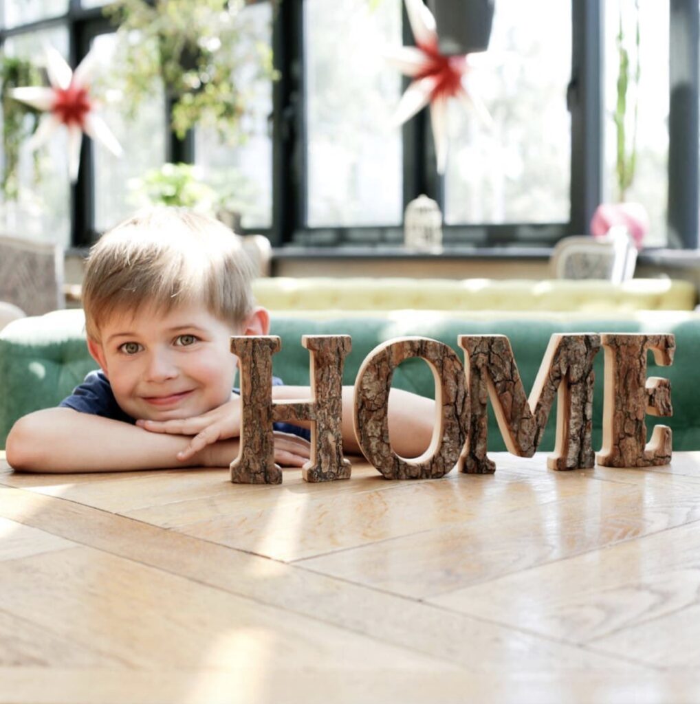 A young boy smiles, resting his chin on a table next to the word "home" spelled out in decorative letters, inside a bright room with windows.