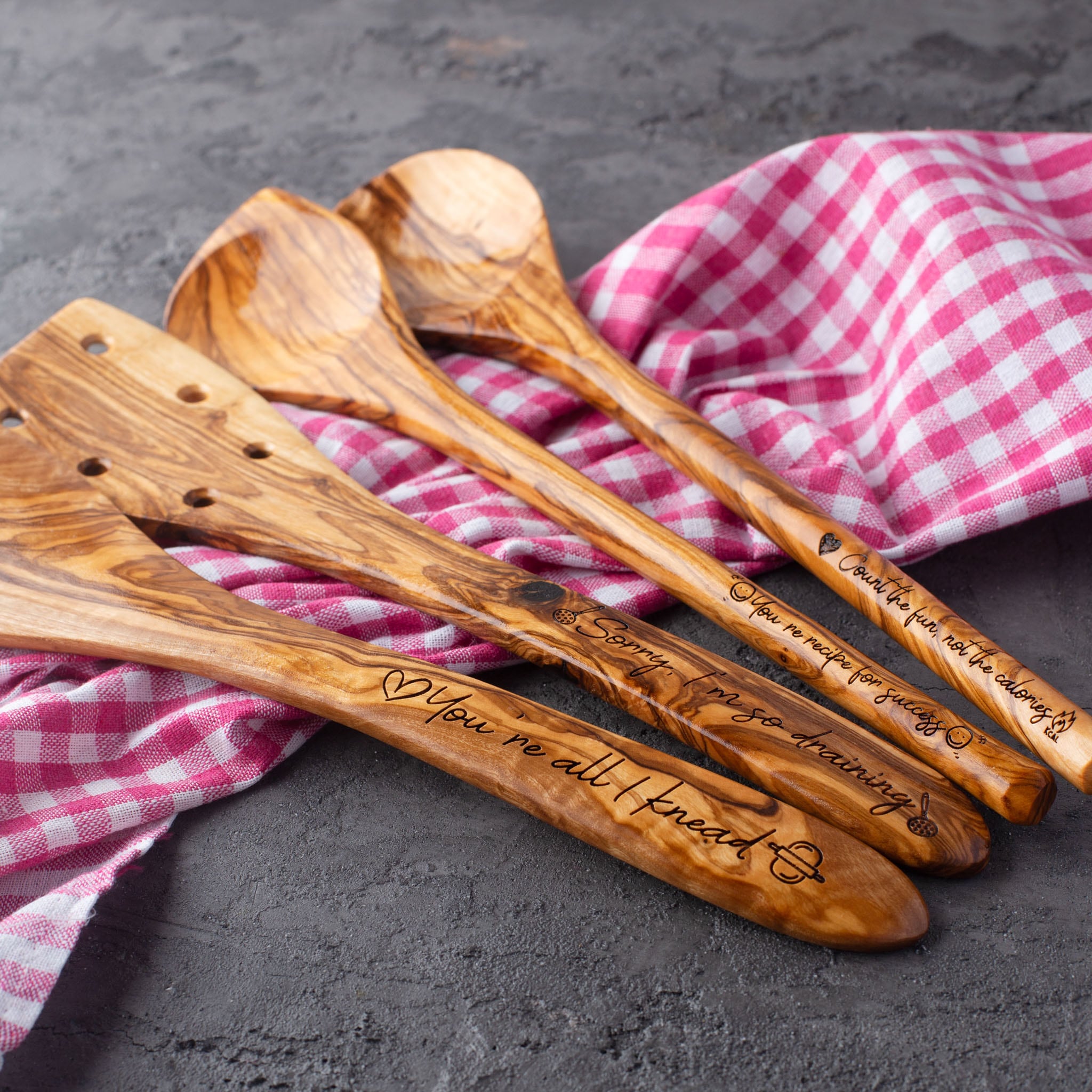 Five wooden cooking spoons with engraved messages, resting on a pink and white checkered cloth on a dark surface.