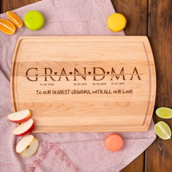 Personalized cutting board with "grandma" carved out, accompanied by dates and a message, flanked by colorful macarons and fruit slices.
