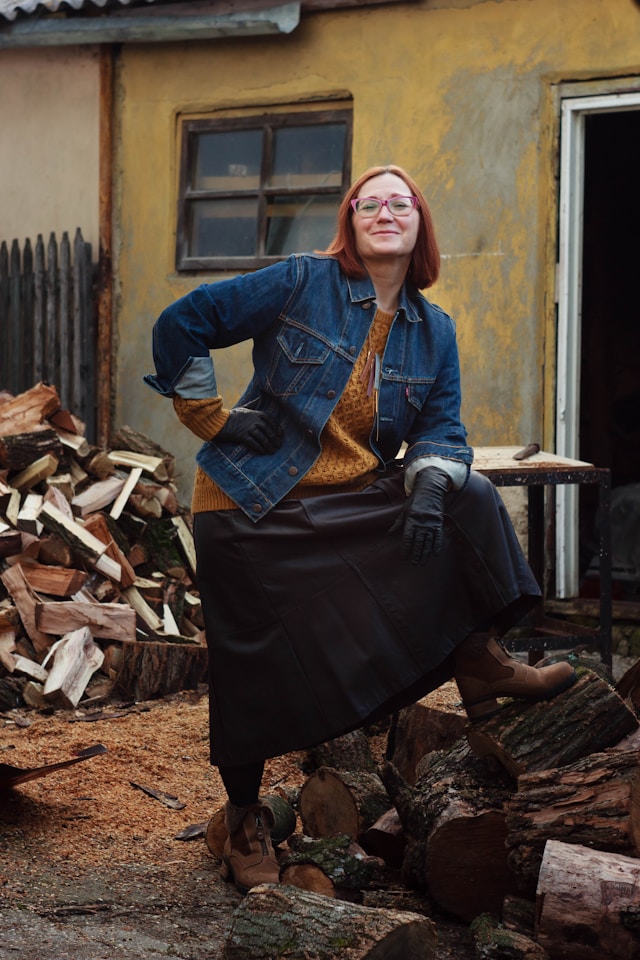 A woman in a denim jacket and black skirt strikes a playful pose beside a stack of firewood, with a rustic yellow house in the background.